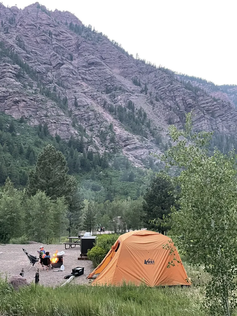 Orange tent with the rain cover and boys sitting in chairs next to a campfire in front of a large rocky mountain