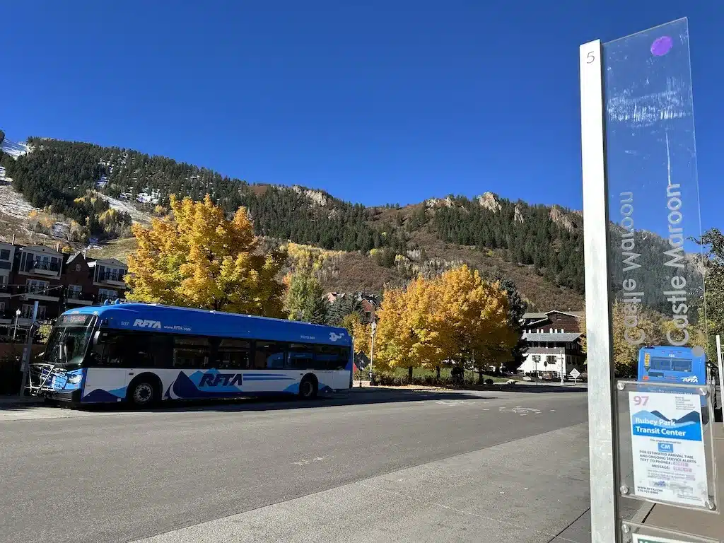 A blue bus parked in front of a mountain covered in fall foliage.