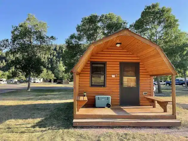 Small wooden cabin in a campground with a porch, steps with trees around it and blue skies