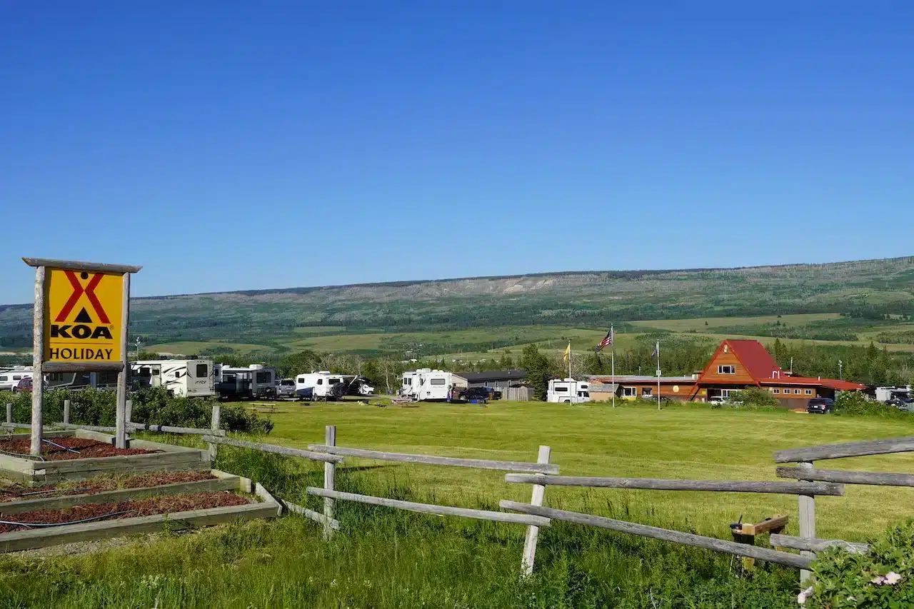 A yellow KOA sign stands at a campground with sprawling green grass and wooden fence with campers and the red KOA building.