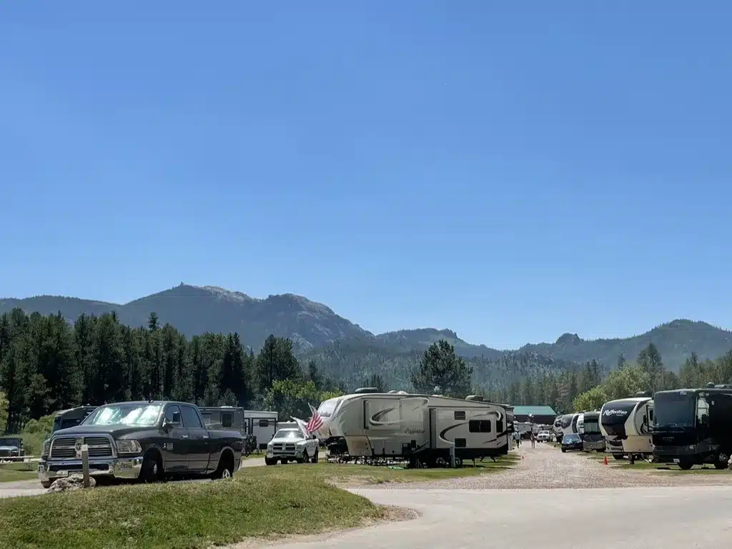夏の青空の下、松林と山々に囲まれたKOAキャンプ場に並ぶRVとトラックの風景