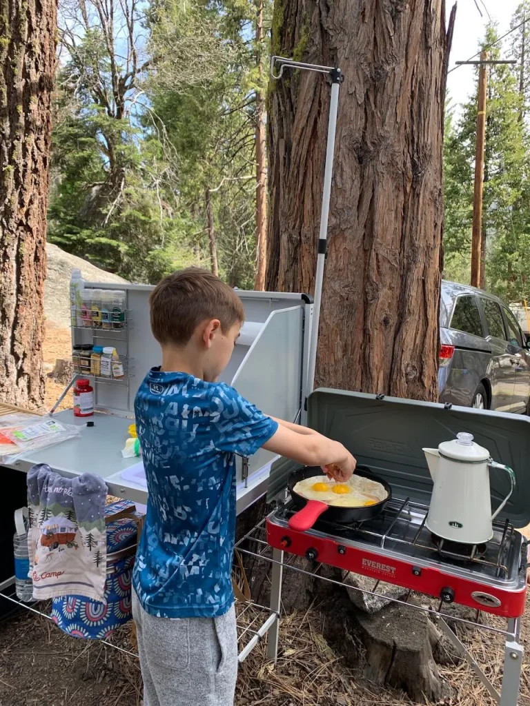 A boy is making breakfast with egg at camp kitchen under the trees at a campsite, example of easy camping meals for family