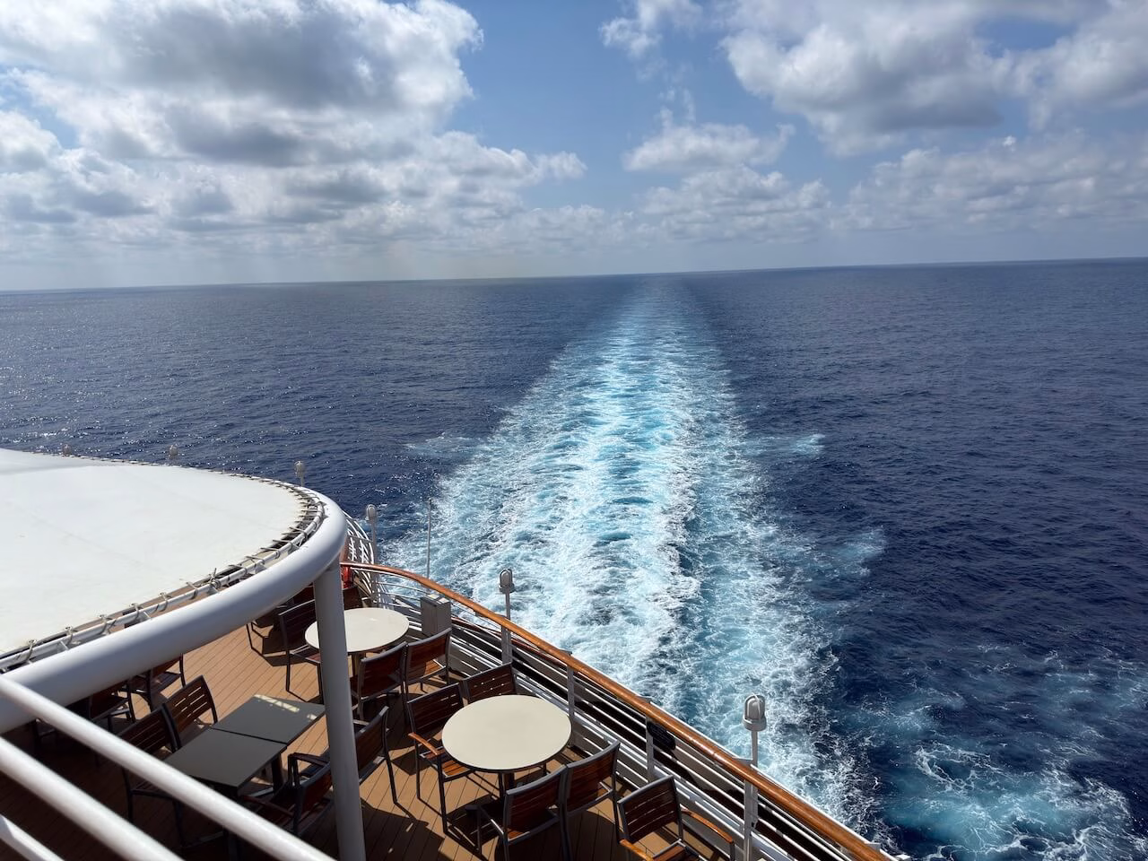 picture of the aft of a cruise ship with tables and chairs on a deck, hand rails and a strait line of the white water behind the ship going off into the distance