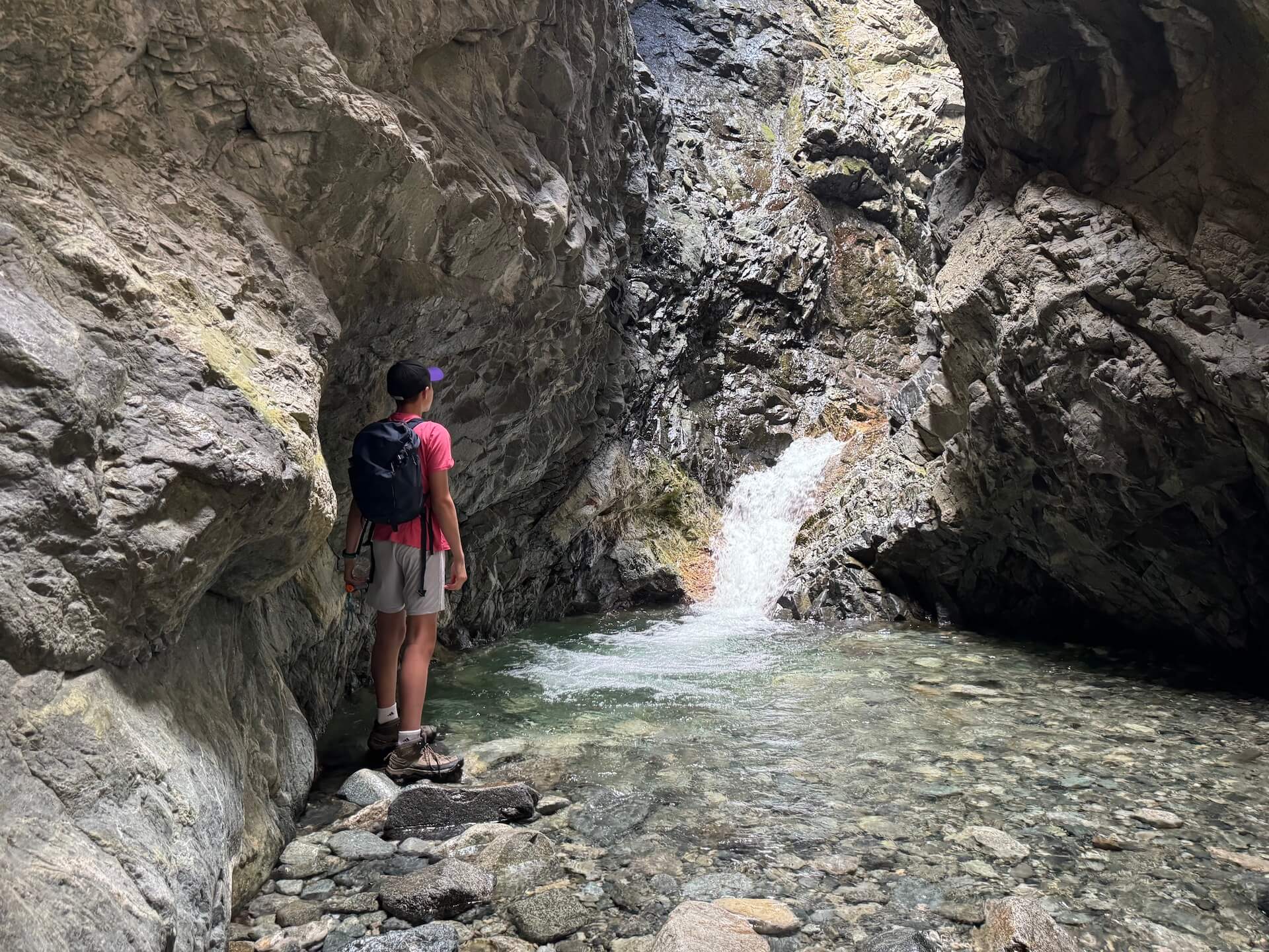 A young boy stands at the water's edge, gazing up at Zapata Falls.