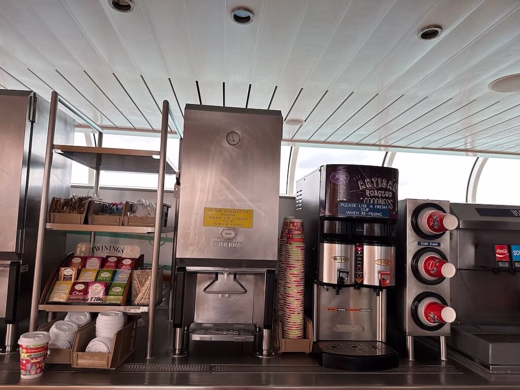 Drink station on a Disney Cruise Line ship offering complimentary soft drinks, soda, coffee, tea, hot chocolate, and water. Guests can enjoy a variety of beverages at this self-service station located on the pool deck.