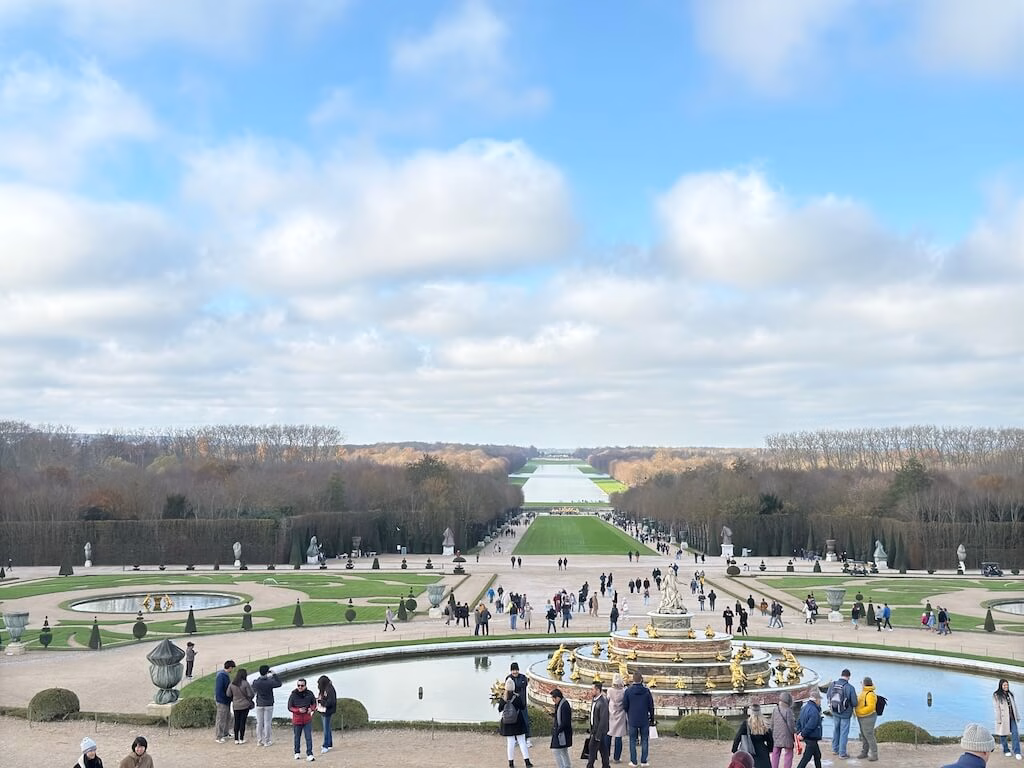 The Versailles gardens, viewed from the palace, are a stunning display of French formal design, featuring symmetrical lawns, vibrant flower beds, sculpted hedges, and shimmering fountains that blend gracefully into the horizon.