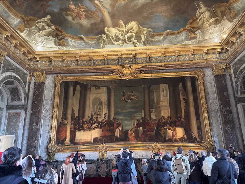 The Hercules Room in the Palace of Versailles is a grand space adorned with opulent marble, intricate gilded details, and a magnificent ceiling painting depicting the Apotheosis of Hercules, showcasing the grandeur of French Baroque artistry
