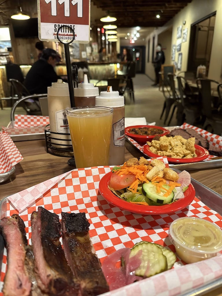 Inside Spitfire Smokehouse in Moab, Utah. A red-checkered tray holds smoked ribs, a fresh salad, pickles, and dipping sauce, served alongside a pint of craft beer. The casual dining area features warm lighting and a laid-back atmosphere. Inside Spitfire Smokehouse in Moab, Utah. A red-checkered tray holds smoked ribs, a fresh salad, pickles, and dipping sauce, served alongside a pint of craft beer. The casual dining area features warm lighting and a laid-back atmosphere. Inside Spitfire Smokehouse in Moab, Utah. A red-checkered tray holds smoked ribs, a fresh salad, pickles, and dipping sauce, served alongside a pint of craft beer. The casual dining area features warm lighting and a laid-back atmosphere.