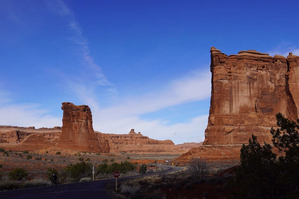Scenic road winding through towering red rock formations in Arches National Park near Moab, Utah, with clear blue skies highlighting the dramatic desert landscape.