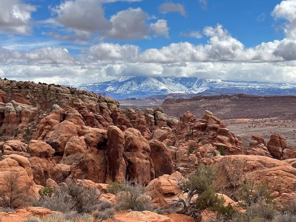 View of red rock formations inside Fiery Furnace at Arches National Park with the snow-capped La Sal Mountains in the distance, showcasing the dramatic scenery on a Moab hiking trail.