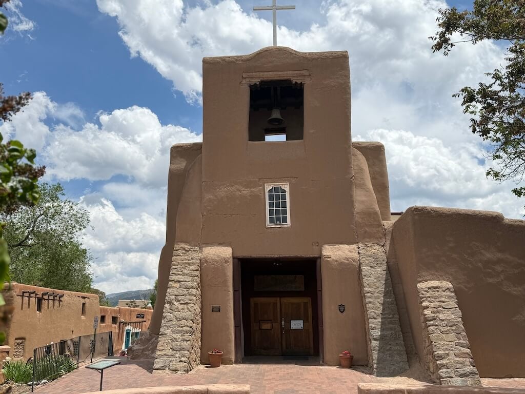San Miguel Chapel – Historic Adobe Church in Santa Fe, New Mexico - Tomo Travel & Camping ニューメキシコ州サンタフェにあるアメリカ最古の教会、サン・ミゲル礼拝堂の正面。アドビ建築と木造の鐘楼が夏空に映える歴史的スポット