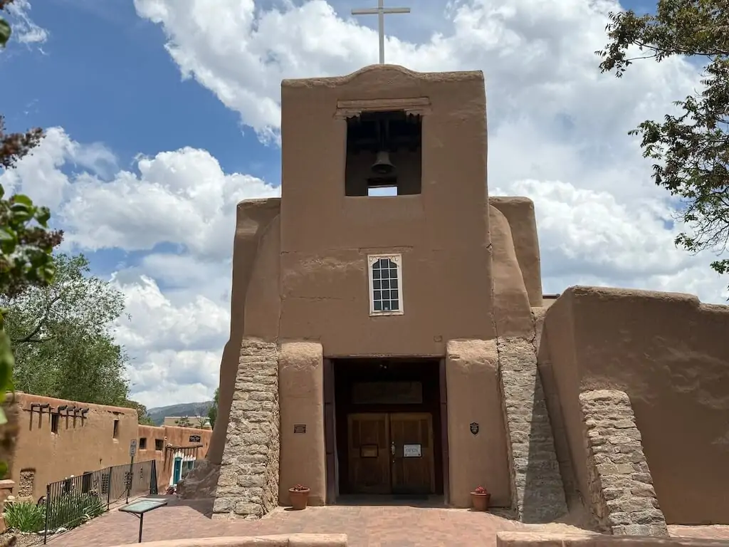 Front view of San Miguel Chapel in Santa Fe, New Mexico—America’s oldest church—featuring traditional adobe architecture and a wooden bell tower under a bright summer sky.