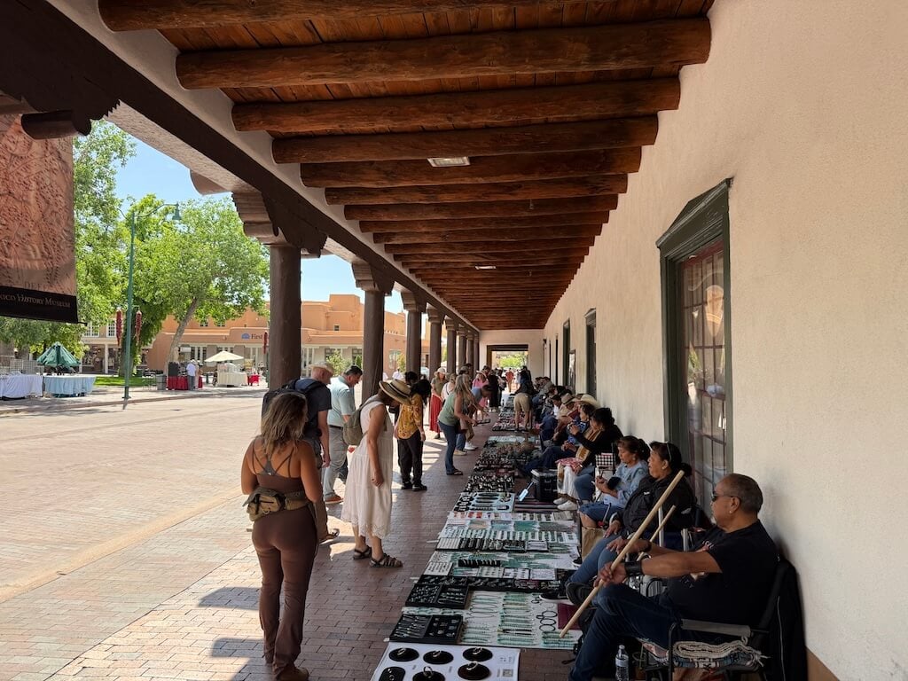 Santa Fe Plaza – A Must-See Cultural Hub and Top Pick for Where to Go in Santa Fe - Tomo Travel & Camping 歴史あるサンタフェ・プラザの屋根付き歩道で、先住民アーティストたちが手作りのネイティブアメリカンジュエリーを販売している様子。本物のローカル文化に触れられる、サンタフェ観光の定番スポット。