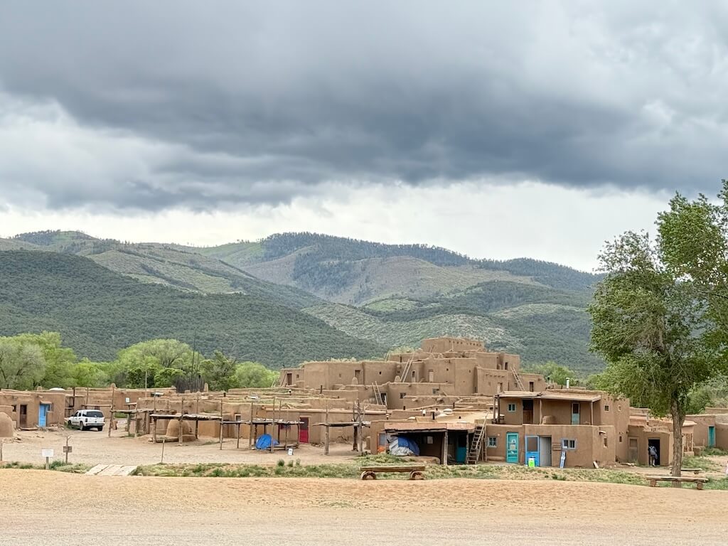 Wide-angle view of Taos Pueblo village with adobe homes and the Sangre de Cristo Mountains in the background under cloudy skies.