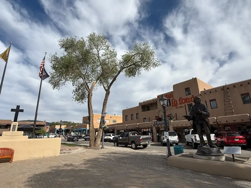 View of Taos Plaza in downtown Taos, New Mexico, featuring the historic Hotel La Fonda and statue under a partly cloudy sky.
