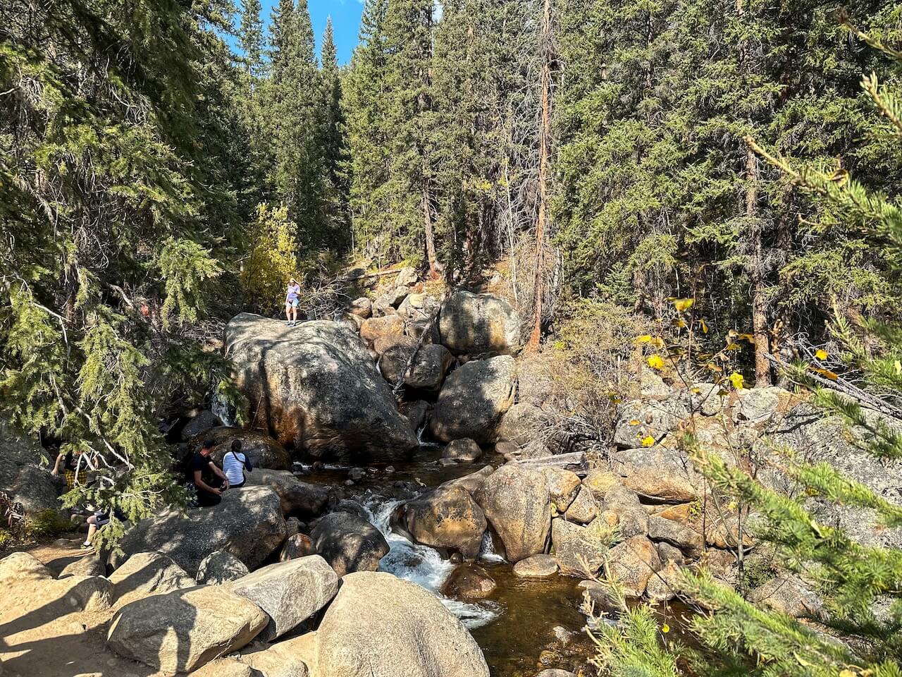 Visitors sitting on large boulders by a mountain creek surrounded by evergreens at Guanella Pass in Colorado.