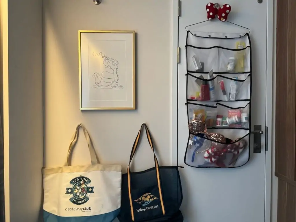 Over-the-door hanging organizer in a Disney Cruise stateroom storing medicine, sunscreen, and Minnie ears with Castaway Club tote bags below.