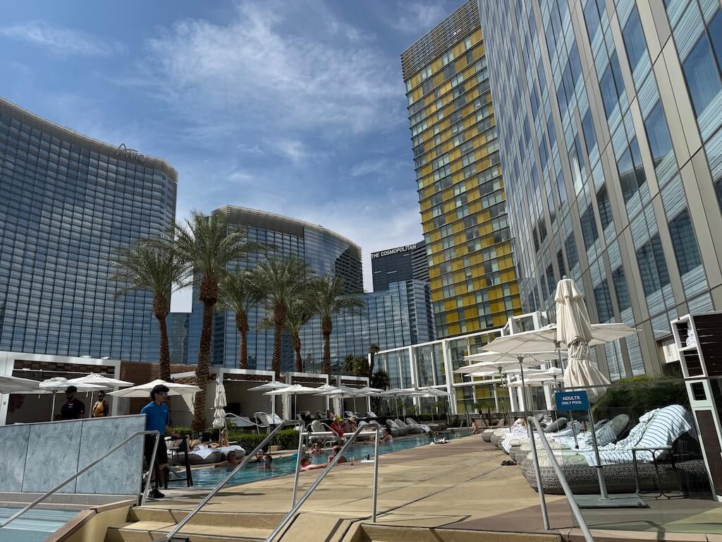 Waldorf Astoria Las Vegas Pool Deck – Adults-Only Luxury Pool with City Views - Tomo Travel & Camping ウォルドルフ・アストリア・ラスベガスの屋外プールエリア。ヤシの木とデッキチェアが並び、ザ・コスモポリタンとアリアを望む眺めが楽しめる。