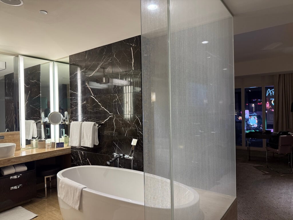 Waldorf Astoria Las Vegas Bathroom – Luxury Soaking Tub and Marble Finishes - Tomo Travel & Camping ウォルドルフ・アストリア・ラスベガスのモダンなバスルーム。深めのバスタブと大理石の壁、ダブルシンクを備え、ラスベガス・ストリップの眺めを楽しめます。