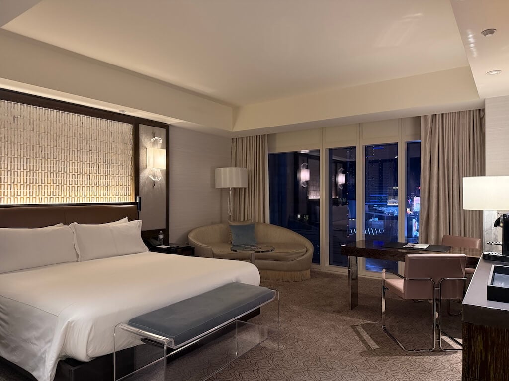 Waldorf Astoria Las Vegas King Room – Luxury Hotel Room with City View - Tomo Travel & Camping ウォルドルフ・アストリア・ラスベガスの広々としたキングルーム。床から天井までの大きな窓とモダンなシーティングエリアを備え、夜のラスベガスの街並みを一望できる。