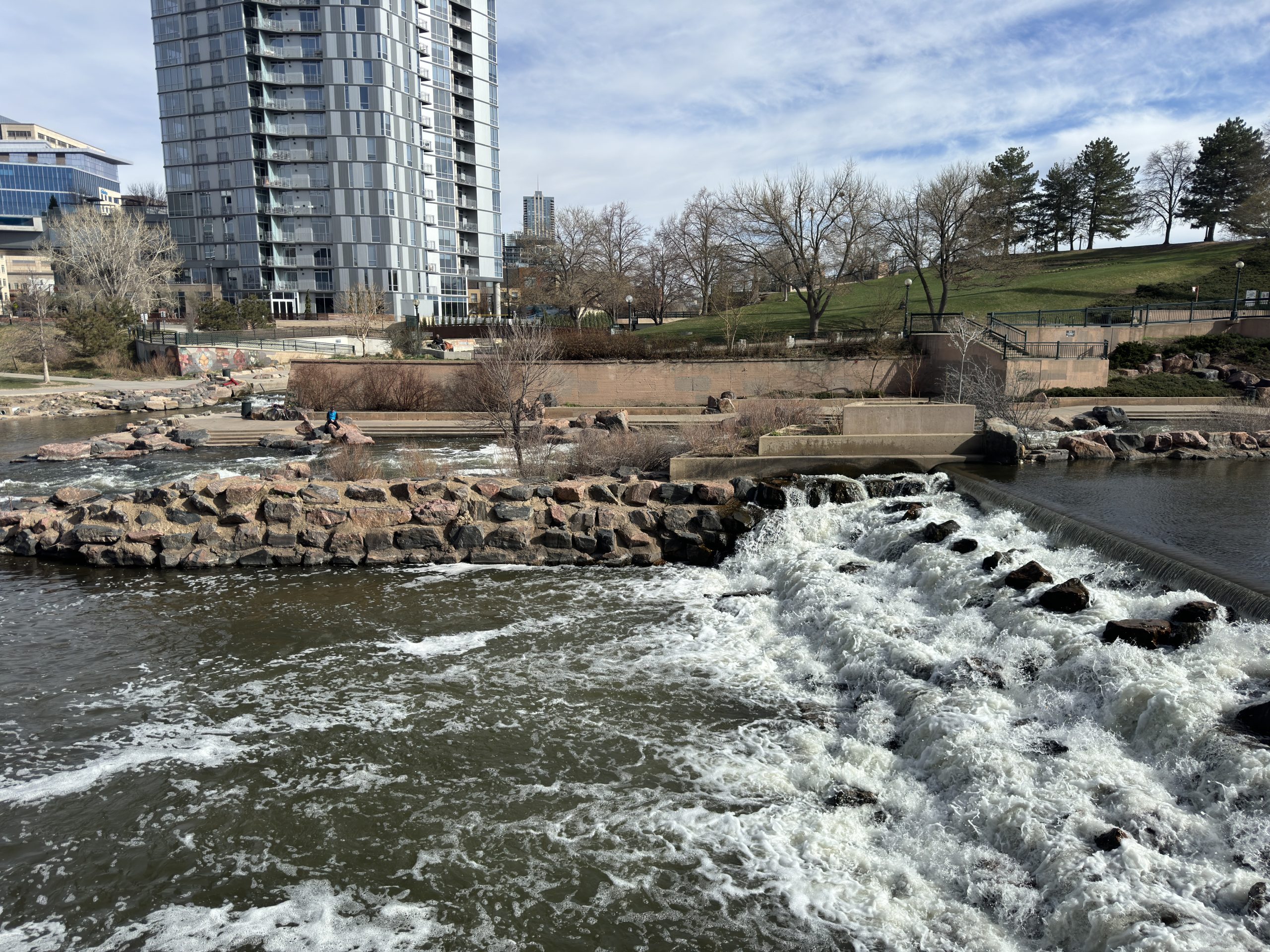 Denver Downtown Confluence Park - Tomo Travel & Camping Riverside park with a stone weir and cascading water, a walkway, and a tall glass apartment building in the background at Confluence Park in downtown Denver.