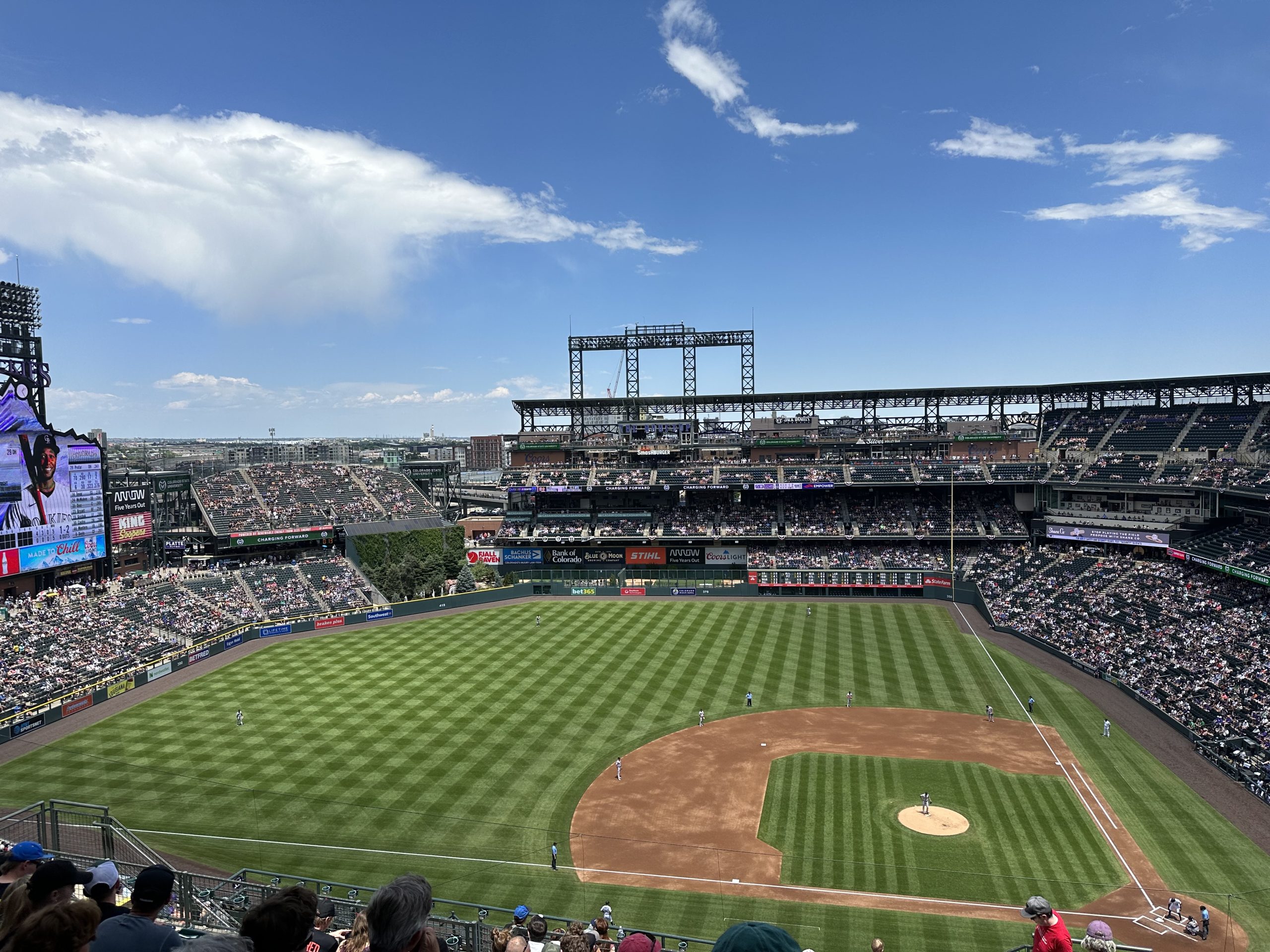 DenverDowntownCoorsFieldRockies - Tomo Travel & Camping A packed baseball stadium with a green of striped infield and pitch, crowds in the stands, and blue sky above at a Colorado Rockies base ball game in Denver, Colorado.