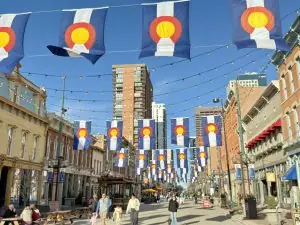 Street scene with Colorado flag bunting overhead, string lights, and pedestrians on a sunny city street lined with shops and buildings from brick to stucco at Larimer Square street with shops and flags in downtown Denver.