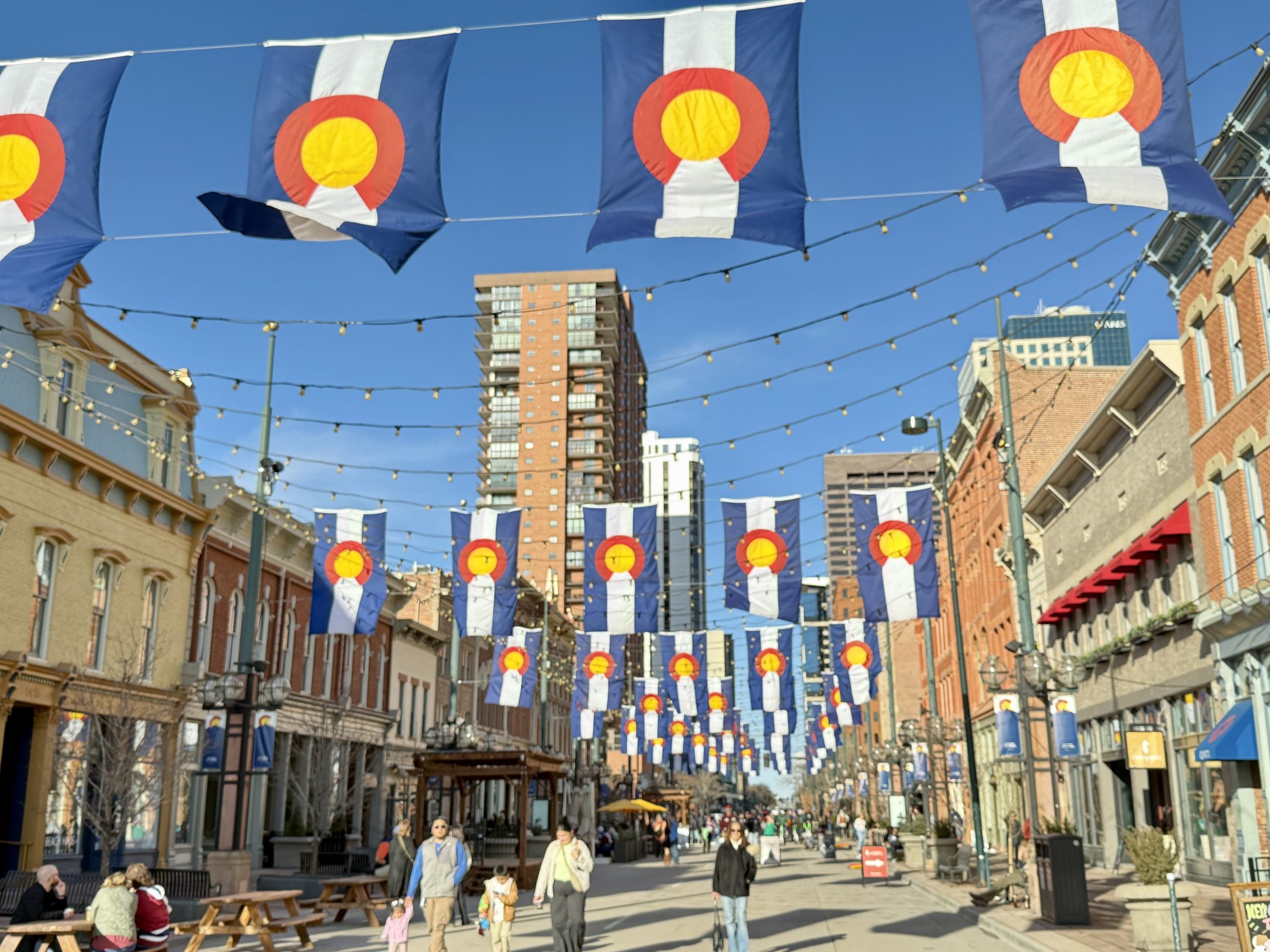 DenverDowntownLarimerSquare - Tomo Travel & Camping Street scene with Colorado flag bunting overhead, string lights, and pedestrians on a sunny city street lined with shops and buildings from brick to stucco at Larimer Square street with shops and flags in downtown Denver.