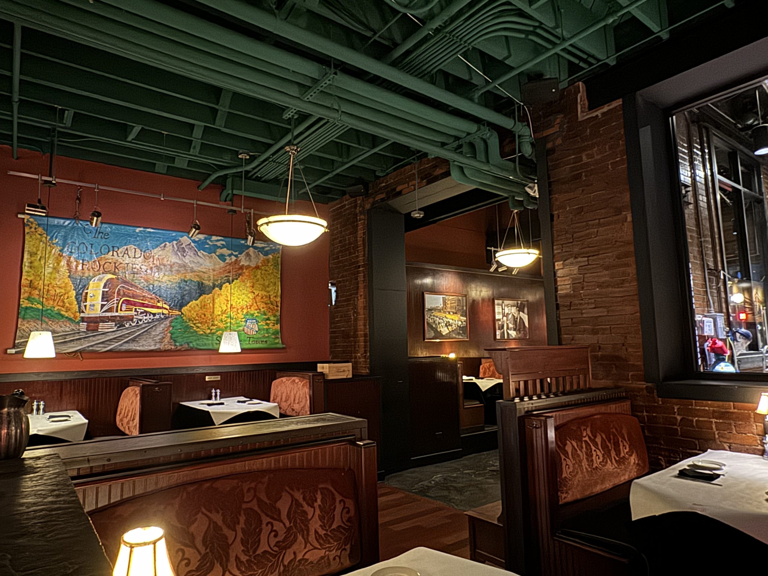 DenverDowntownModernAmerican - Tomo Travel & Camping Restaurant dining area with brick walls, green ceiling pipes, booth seating, and a colorful mural of a train through mountains at Modern American dining setting in a downtown Denver restaurant.