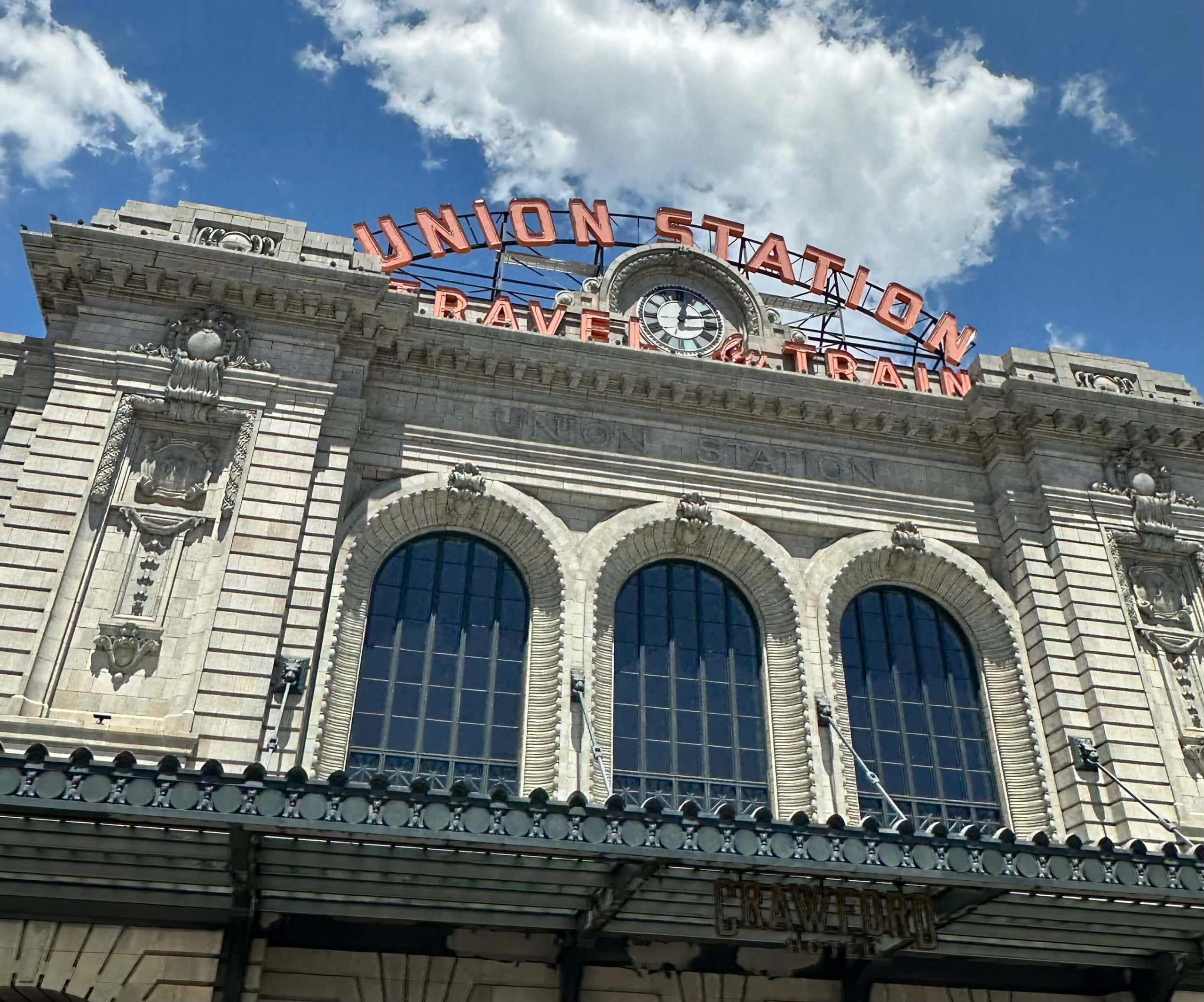 DenverDowntownUnionStation - Tomo Travel & Camping Facade of a historic train station with a large arched window, ornate stonework, and a red'Union Station' sign atop the building against a blue sky at Union Station in Downtown Denver.