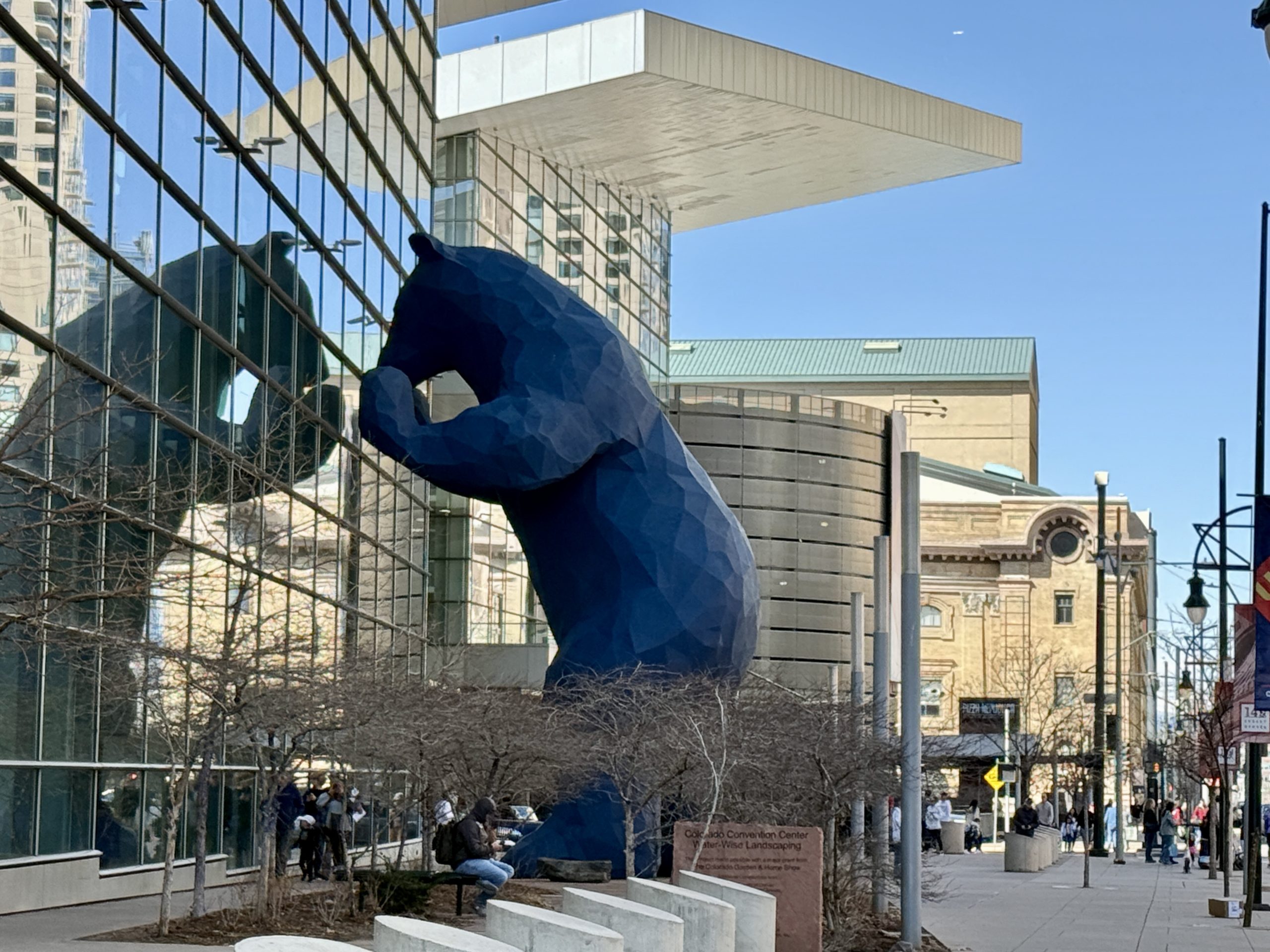 Denver Downtown Big Blue Bear - Tomo Travel & Camping Large dark blue geometric bear sculpture leaning against a glass-fronted building in a city plaza in Denver Downtown.