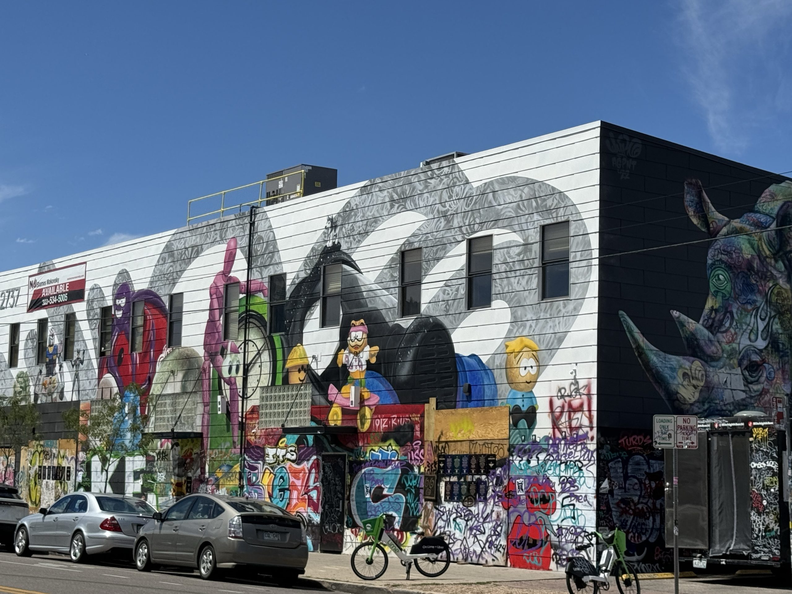 Denver downtown Rino - Tomo Travel & Camping Wide mural on a building wall features colorful cartoon characters and graffiti, with parked cars and a cyclist in front under a clear blue sky; the artwork spans the entire side of the block in the RiNo Art District in Denver.