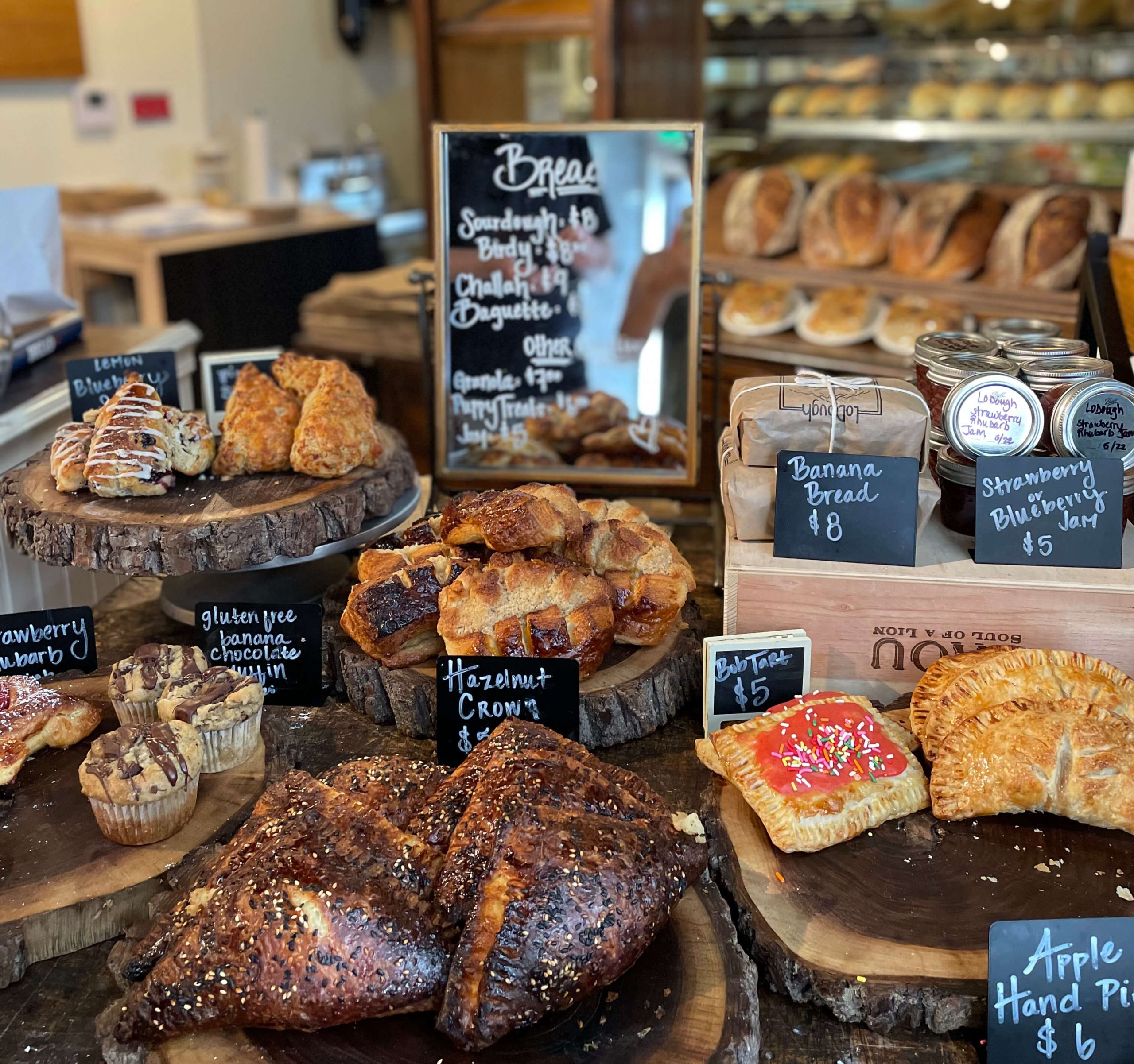 LoDough Bakery Denver - Tomo Travel & Camping Assorted bakery display with sesame-topped loaves, croissants, and pastries on wooden boards and chalkboard signs behind glass at LoDough Bakery in downtown Denver.