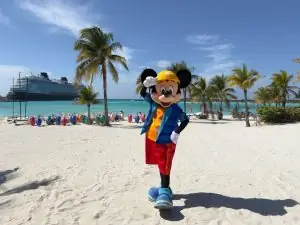 Mickey Mouse wearing a tropical vacation outfit on the white sand beach of Castaway Cay with a Disney Cruise ship in the background.