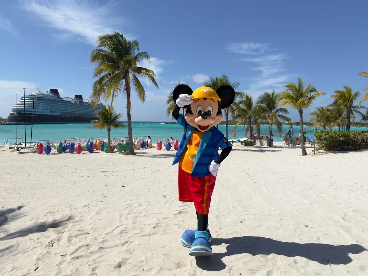 Mickey Mouse wearing a tropical vacation outfit on the white sand beach of Castaway Cay with a Disney Cruise ship in the background.