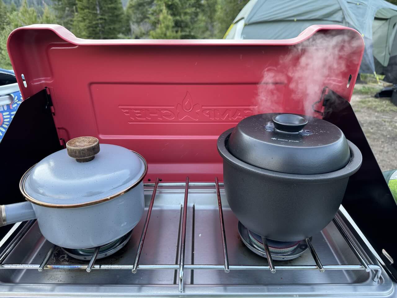 Two Burner Camp Stove Cooking a Family Meal While Tent Camping in a National Park - Tomo Travel & Camping Two pots cooking simultaneously on a red two-burner Camp Chef camp stove with steam rising, a green tent and pine trees visible in the background at a national park campground