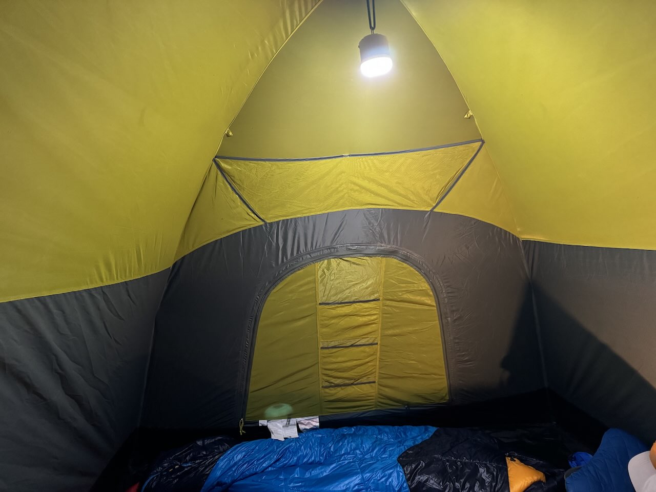 Inside a Family Tent at Night: Tent Camping Light and Sleeping Bags in a National Park - Tomo Travel & Camping Inside view of a green and gray family camping tent at night with a hanging tent light illuminating the interior and blue sleeping bags on the floor