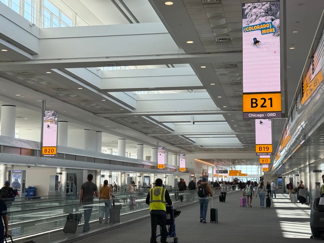DIA Denver Airport - Tomo Travel & Camping Busy airport terminal with moving walkways and hanging B gate signs (B21, B20) in view.