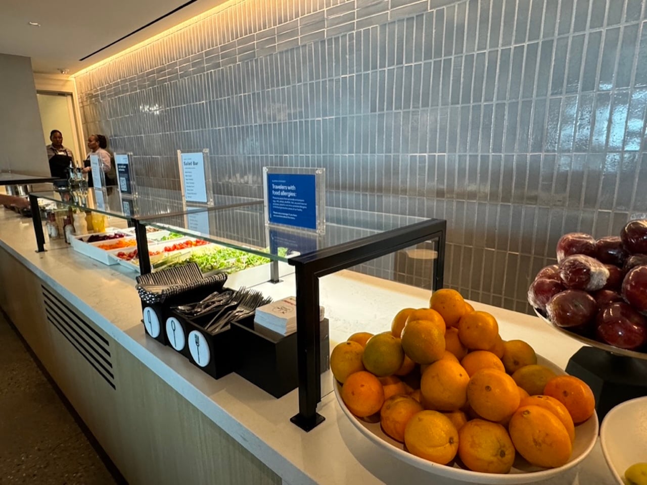 DIA United Club Food - Tomo Travel & Camping Buffet-style salad bar with fresh veggies behind a glass sneeze guard and bowls of citrus fruits at the counter. On the side, a fruit bowl with oranges and plums at United Club Lounge, Denver Airport