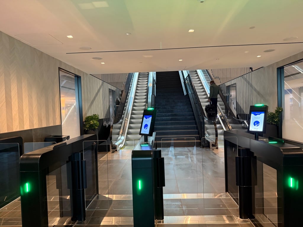 Untied ClubEntrance - Tomo Travel & Camping Modern indoor lobby with glass security gates and green LED lights; escalators rise in the background as a traveler uses the right-side escalator.