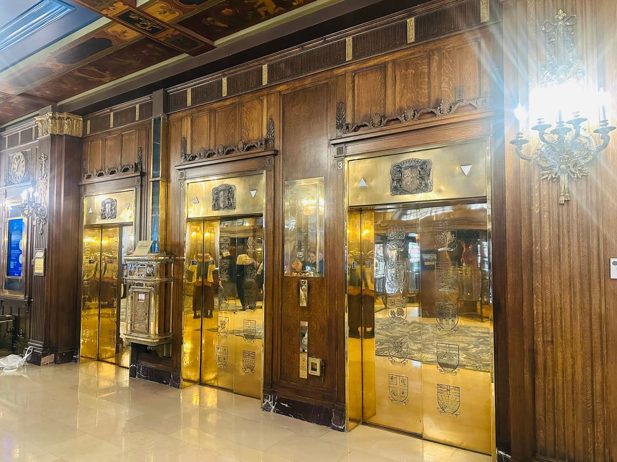 chateau-frontenac-elevators-mailbox - Tomo Travel & Camping Grand hotel lobby with three gold elevator doors set in dark wood paneling and crest emblems, marble base, and reflections in the doors.