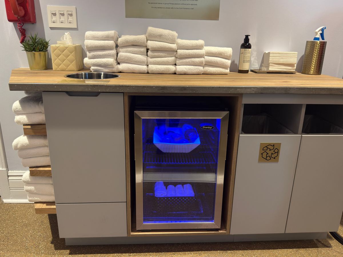 chateau-frontenac-gym-towels - Tomo Travel & Camping Countertop spa/kitchen area with a blue-lit mini fridge in the center, stacked white towels, and assorted supplies on the right (soap, spray bottle, napkins).