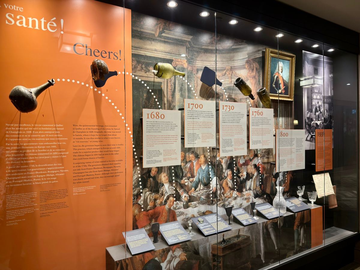 chateau-frontenac-history-exhibit - Tomo Travel & Camping Museum display about wine history with an orange panel reading'Cheers!', timeline cards, and bottles suspended above a historic mural in a glass case.