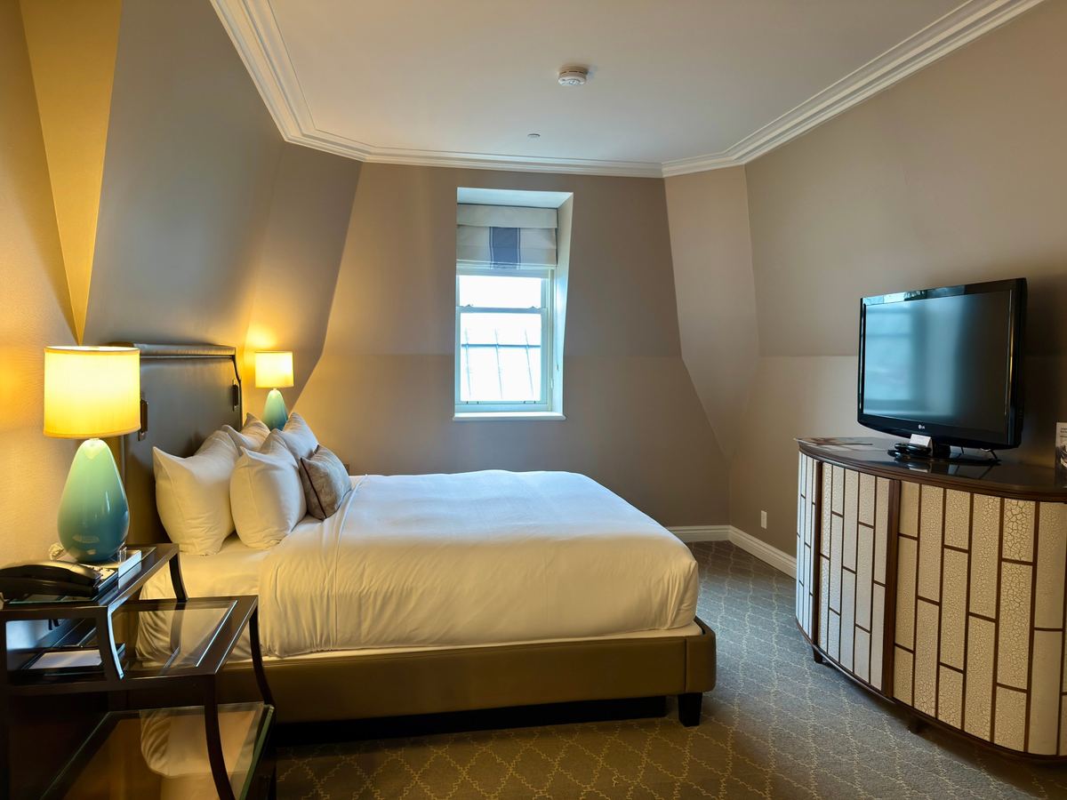chateau-frontenac-king-room - Tomo Travel & Camping Hotel room with a king bed, two teal lamps, window, and a curved cabinet with a flatscreen TV.