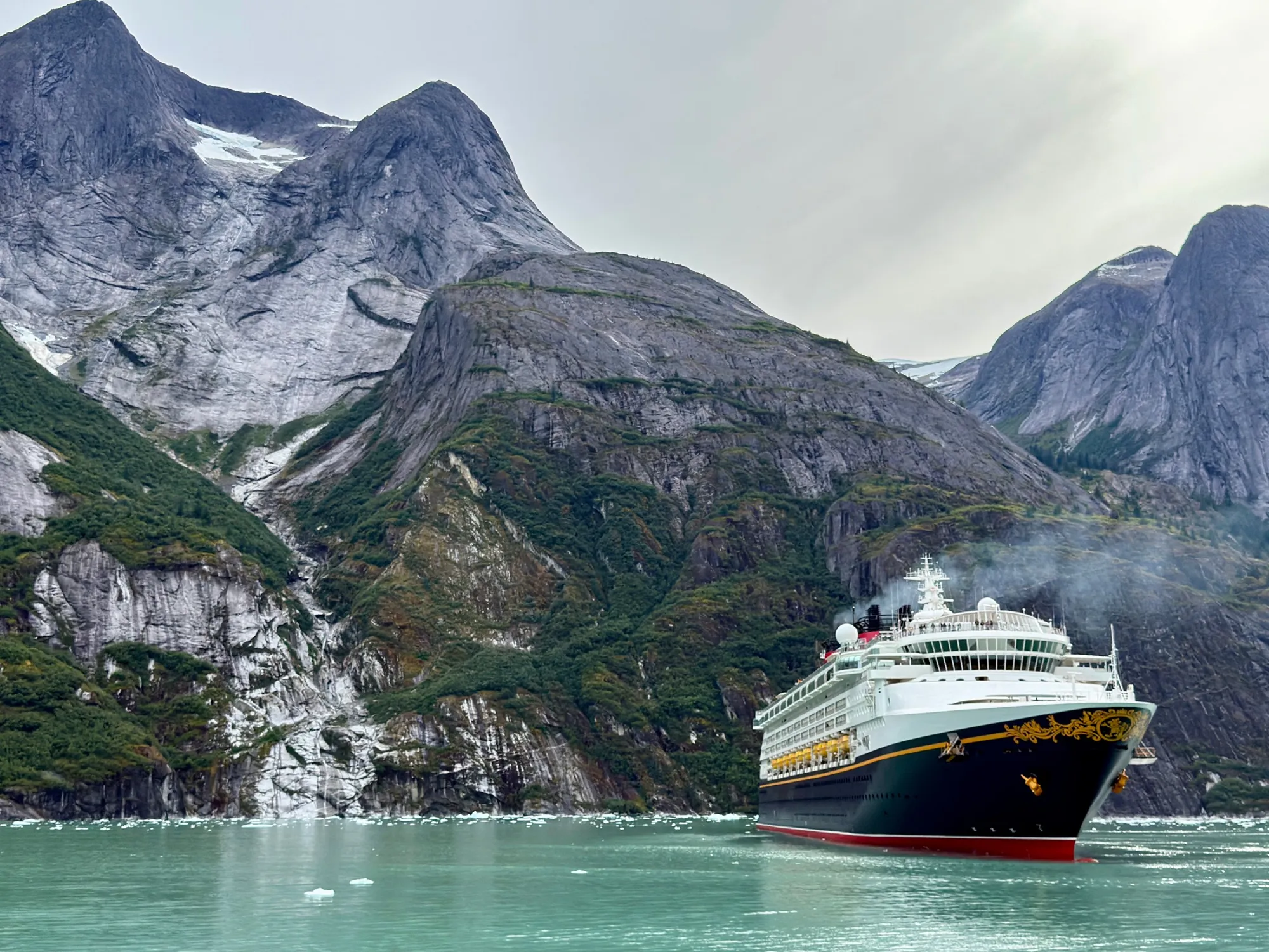 Disney Wonder sailing past steep granite cliffs in an Alaska fjord with floating ice