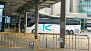 White coach bus parked at a terminal behind yellow metal barriers, with a modern glass building in the background.